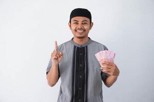 leende eller Lycklig ung asiatisk muslim man finger pekande upp innehav papper pengar gåva thr från familj ramadan eid al-fitr tid bär grå muslim kläder isolerat på vit bakgrund foto