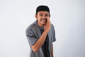 ung asiatisk muslim man bär grå muslim trasa, hand bredvid mun viskande hemligheter eller skvaller isolerat på vit bakgrund foto