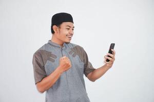 upphetsad eller Lycklig ung asiatisk muslim man innehav på mobil telefon och händer i nävar håller på med vinnare gest bär koko kläder isolerat över vit bakgrund foto