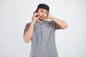 förvirrad ung asiatisk muslim man talande på mobil telefon med händer innehav huvud bär grå muslim kläder isolerat på vit bakgrund foto