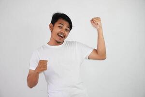 självsäker glad stilig asiatisk man lyft händer upp bär vit t skjorta vinnare gest knäppning nävar. känner lycka visa näve upp Framgång isolerat på vit bakgrund foto