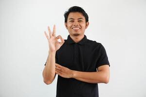 ung asiatisk man leende vänlig medan ger ok finger tecken bär svart polo t skjorta isolerat på vit bakgrund foto