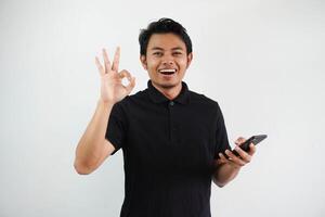 ung asiatisk man leende självsäker och ge ok finger tecken medan innehav en mobil telefon bär svart polo t skjorta isolerat på vit bakgrund foto