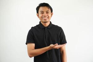 leende ung asiatisk man i svart polo skjorta, vit bakgrund som visar en kopia Plats på en handflatan och innehav annan hand på midja. foto