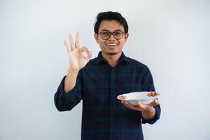 ung asiatisk man leende Lycklig och ge ok tecken med hand medan som visar tömma middag tallrik isolerat på vit bakgrund. foto