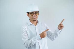 ung asiatisk arkitekt man bär vit hård hatt säkerhet hjälm ser kamera med finger pekande de vänster sida isolerat på vit bakgrund foto