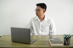 ung asiatisk affärsman i en arbetsplats framställning tvivel gest ser sida, bär vit skjorta med glasögon isolerat foto