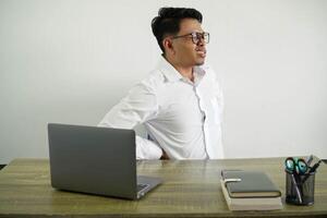 ung asiatisk affärsman i en arbetsplats lidande från ryggvärk för har tillverkad ett ansträngning ha på sig vit skjorta med glasögon isolerat foto