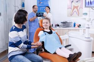 Lycklig liten flicka bär dental haklapp i tandläkare kontor innehav förälder hand väntar tand undersökning. barn med henne mor under tänder kolla upp upp med stomatolog Sammanträde på stol. foto