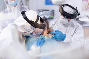 caucasian liten flicka har dental behandling på tandläkarens kontor Sammanträde på stol med haklapp. stomatologi team bär ppe kostym under covid19 håller på med procedur på barn tänder. foto