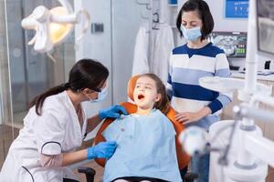 tandläkare håller på med tänder kolla upp upp av liten flicka Sammanträde på dental stol bär haklapp. tandvård specialist under barn hålighet samråd i stomatologi kontor använder sig av modern teknologi. foto