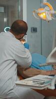 tandläkare tekniker med handskar förklara de kirurgi belysning de lampa. ortodontist tala till kvinna med tandvärk Sammanträde på stomatologisk stol medan sjuksköterska framställning verktyg för undersökning. foto