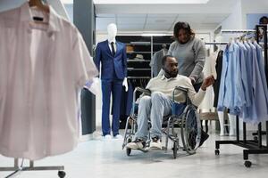 afrikansk amerikan man i rullstol välja kläder medan handla med flickvän i Kläder Lagra. hackning Centrum kund med kronisk nedsättning granskning tillfällig shirts på kuggstång foto