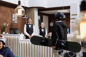 manlig besökare ankommer med resväska och åka snowboard redskap på åka skidor hotell lobby för checka in på disken. vänlig anställda på främre skrivbord bistå gäst med bokning på vinter- berg tillflykt. foto