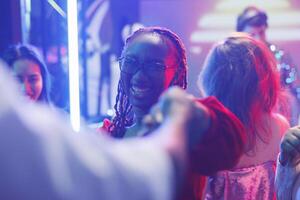 Lycklig kvinna dans med vänner och festa på diskotek i nattklubb. glad leende ung afrikansk amerikan clubber rör på sig på dansgolv på elektronisk musik prestanda foto