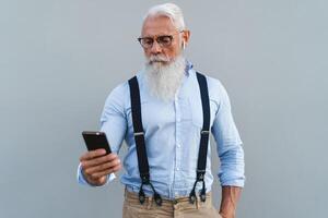 senior man använder sig av mobil smartphone och lyssnande musik med trådlös hörlurar - mode äldre manlig arbetssätt med teknologi enheter foto