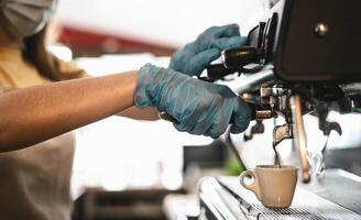 ung kvinna sida se framställning kaffe espresso medan bär kirurgisk mask och handskar för förebyggande korona virus spridning - bar ägare säkerhet arbetssätt - varm drycker och covid-19 regler begrepp foto