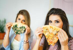 ung skön kvinnor äter skivor av smak italiensk pizza på Hem - Lycklig Söt systrar beläggning deras ansikten med snabb mat i lägenhet - begrepp av människor, livsstil, måltid - fokus på kvinna öga foto