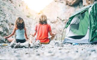 hälsa ung par håller på med yoga Nästa till brand medan camping med tält på en berg - vänner mediterar tillsammans på stenar på solnedgång - människor, hälsa, livsstil och sporter begrepp foto