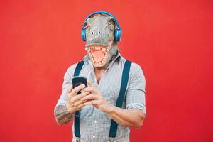 senior mode man bär t Rex mask använder sig av mobil smartphone lyssnande musik med hörlurar - galen bisarr hipster kille har roligt med ny teknologi - rolig och absurd overkligt begrepp foto