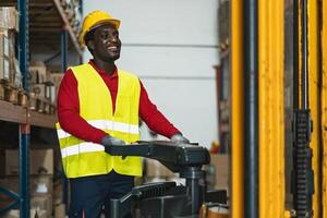 svart man arbetssätt i lager med gaffeltruck läser in leverans lådor - logistisk och industri begrepp foto