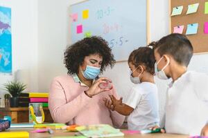 lärare rengöring händer till studerande barn med desinfektionsmedel gel medan bär ansikte mask i förskola klassrum under korona virus pandemi - sjukvård och utbildning begrepp foto
