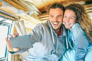 Lycklig man och kvinna tar en selfie med en mobil smart telefon kamera - resa par framställning bilder av deras resa på en årgång minibuss med trä interiör - resa, kärlek, teknologi begrepp foto