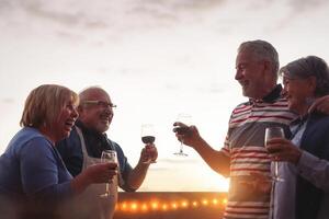 Lycklig senior vänner dricka röd vin på utegrill middag i terrass - mogna människor glädjande och skrattande tillsammans på solnedgång på taket - vänskap och äldre pensionering livsstil begrepp foto