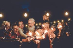 Lycklig familj har på middag fest utomhus- - grupp av raser äldre och ung människor fira tillsammans dricka vin innehav fyrverkeri sparklers - begrepp av ungdom och äldre föräldraskap foto