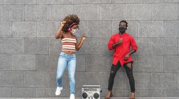 ung afrikansk människor bär ansikte mask dans utomhus- medan lyssnande till musik med trådlös hörlurar och årgång boombox foto