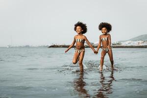 Lycklig systrar löpning tillsammans inuti vatten under sommar tid - afro barn har roligt spelar på de strand - familj kärlek och resa semester livsstil begrepp foto