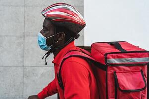 afrikansk ryttare man leverera måltid till kunder med elektrisk cykel medan bär ansikte mask under korona virus utbrott - eco snabb leverans mat begrepp foto