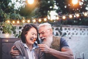 Lycklig senior par har roligt på middag hus fest - äldre människor med annorlunda etnicitet håller på med en romantisk datum för fira årsdag - äldre, mat, dryck och kärlek begrepp foto