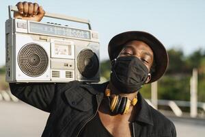 ung afrikansk man lyssnande till musik med årgång boombox stereo medan bär ansikte mask utomhus- foto