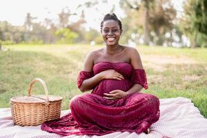 afrikansk kvinna smeksam henne gravid mage medan håller på med en picknick i parkera - moderskap livsstil begrepp foto