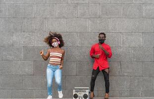 ung afrikansk människor bär ansikte mask och dans utomhus- medan lyssnande till musik med trådlös hörlurar och årgång boombox foto