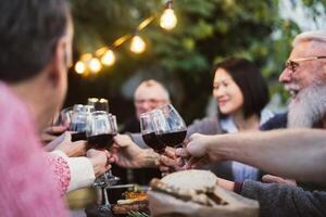 Lycklig familj dining och provsmakning röd vin glasögon i utegrill middag fest - människor med annorlunda åldrar och etnicitet har roligt tillsammans foto