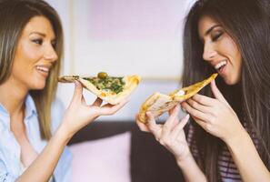 ung skön kvinnor äter skivor av gott italiensk pizza på Hem - Lycklig Söt damer njuter en snabbt måltid tillsammans i deras lägenhet - begrepp av människor, livsstil, mat - fokus på pizza foto