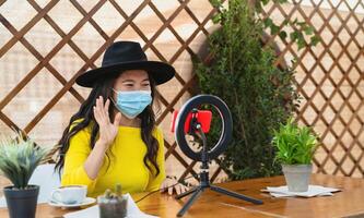 Lycklig asiatisk kvinna bär ansikte mask och vlogging uppkopplad använder sig av smartphone kam och influencer led medan dricka en kaffe Sammanträde i bar under korona virus utbrott foto