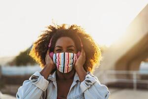 Lycklig afro kvinna lyssnande till Spellista musik med trådlös hörlurar medan bär ansikte färgad mask utomhus- foto