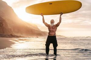 Lycklig passa senior har roligt surfing på solnedgång tid - sportig skäggig man Träning med surfingbräda på de strand - äldre friska människor livsstil och extrem sport begrepp foto