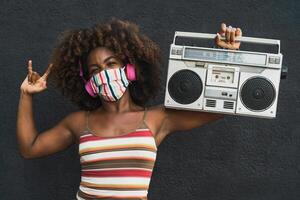 ung afro kvinna bär ansikte mask dans utomhus- medan lyssnande till musik med trådlös hörlurar och årgång boombox stereo foto