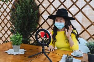 Lycklig asiatisk kvinna bär ansikte mask och vlogging uppkopplad använder sig av smartphone kam och influencer led medan dricka en kaffe Sammanträde i bar under korona virus utbrott foto