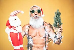 Lycklig passa santa claus utrustade med vit jul lampor - trendig skägg hipster senior innehav en mini santa claus marionett och xmas träd - firande och högtider begrepp foto