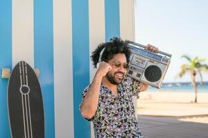 Lycklig afro man har roligt lyssnande till musik med trådlös hörlurar och årgång boombox på de strand under sommar tid foto