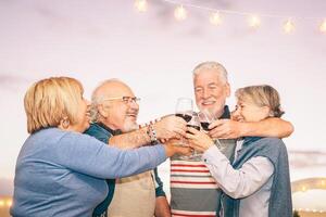 Lycklig senior vänner glädjande och toasting med röd vin på terrass - äldre människor har roligt på middag fest på uteplats på solnedgång - vänskap, dryck och äldre pensionering livsstil begrepp foto