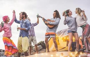 grupp vänner håller på med fest spelar gitarr och camping utomhus- - Lycklig ung människor har roligt dricka öl och skrattande i läger by - ungdom kultur livsstil och semester resa begrepp foto