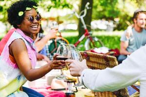 ung människor har en underbar picknick i en parkera njuter de glad ögonblick dricka och äter. Lycklig vänner toasting glasögon av vin. afro hipster kvinna har en Skål med henne vänner foto