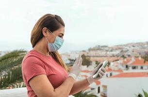 mogna latin kvinna bär kirurgisk ansikte mask och handskar använder sig av mobil smartphone för video kallelse på Hem terrass - sjukvård covid19 förebyggande och teknologi begrepp foto