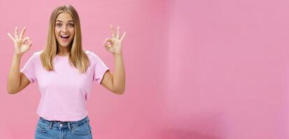porträtt av entusiastisk attraktiv caucasian flicka i trendig t-shirt och jeans som visar Okej eller bekräfta gest med roade bred leende stående nöjd över rosa bakgrund reagerar till excellent Nyheter foto
